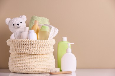 Photo of Knitted basket with baby cosmetic products, bath accessories and toy bear on white table against beige background. Space for text