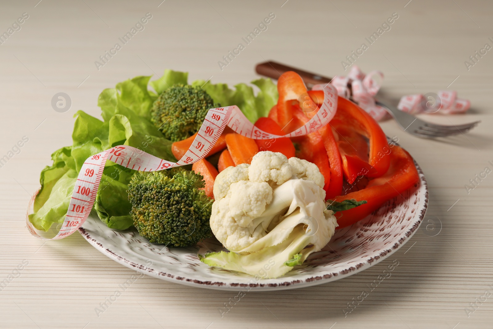 Photo of Healthy diet. Plate of fresh vegetables, measuring tape and fork on light wooden table