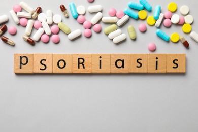 Photo of Word Psoriasis made of wooden squares with letters and different pills on light grey background, flat lay. Space for text