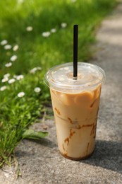 Takeaway plastic cup with cold coffee drink and straw near green grass outdoors