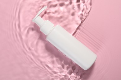 Photo of Bottle of face cleansing product in water against pink background, top view