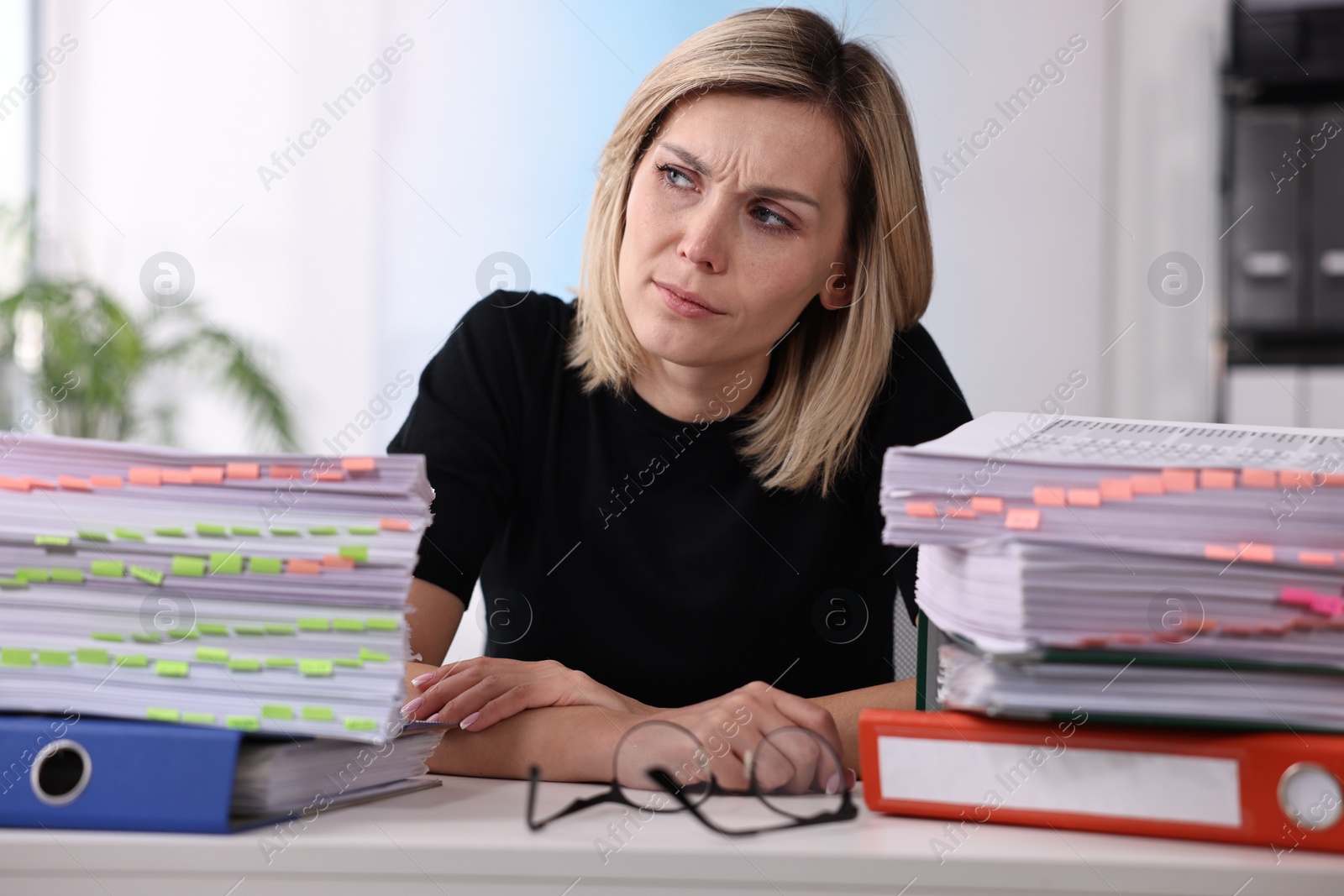 Photo of Overwhelmed woman sitting at table with stacks of documents and folders in office
