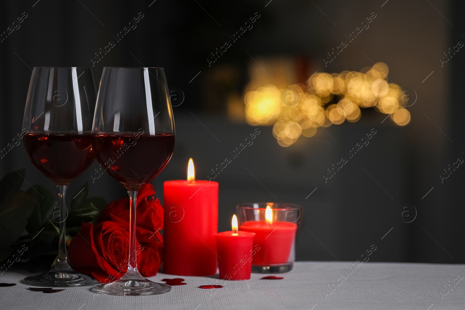 Photo of Glasses of red wine, burning candles and rose flowers on grey table against blurred lights, space for text. Romantic atmosphere