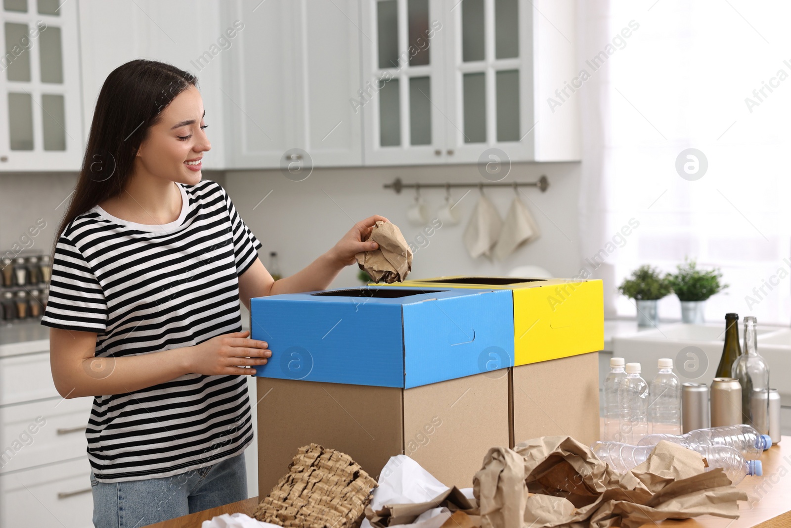 Photo of Garbage sorting. Smiling woman throwing crumpled paper into cardboard box in kitchen