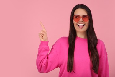 Photo of Happy young woman with heart shaped glasses showing her tongue and pointing on pink background. Space for text