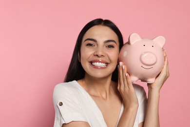 Emotional young woman with ceramic piggy bank on pale pink background