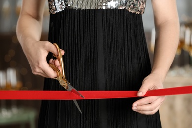 Woman cutting red ribbon with scissors indoors at ceremonial event