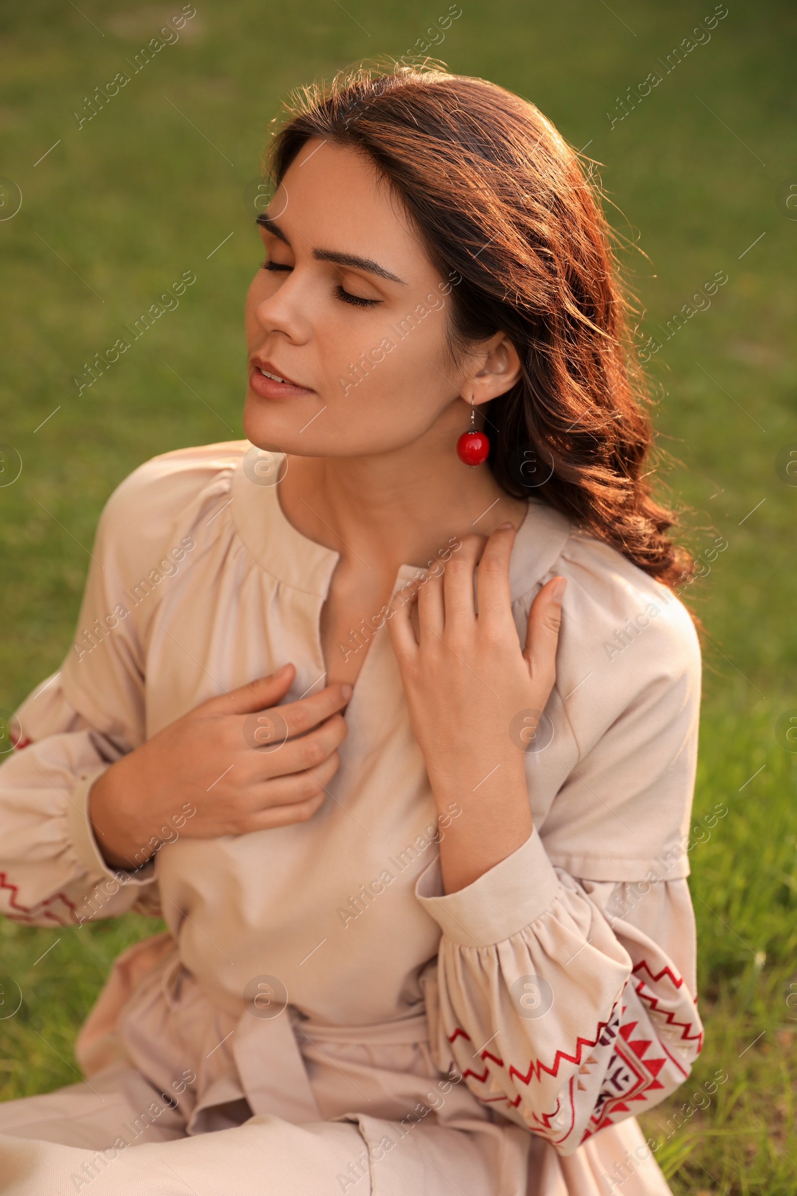 Photo of Beautiful woman in embroidered dress sitting on green grass outdoors. Ukrainian national clothes