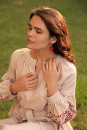 Photo of Beautiful woman in embroidered dress sitting on green grass outdoors. Ukrainian national clothes