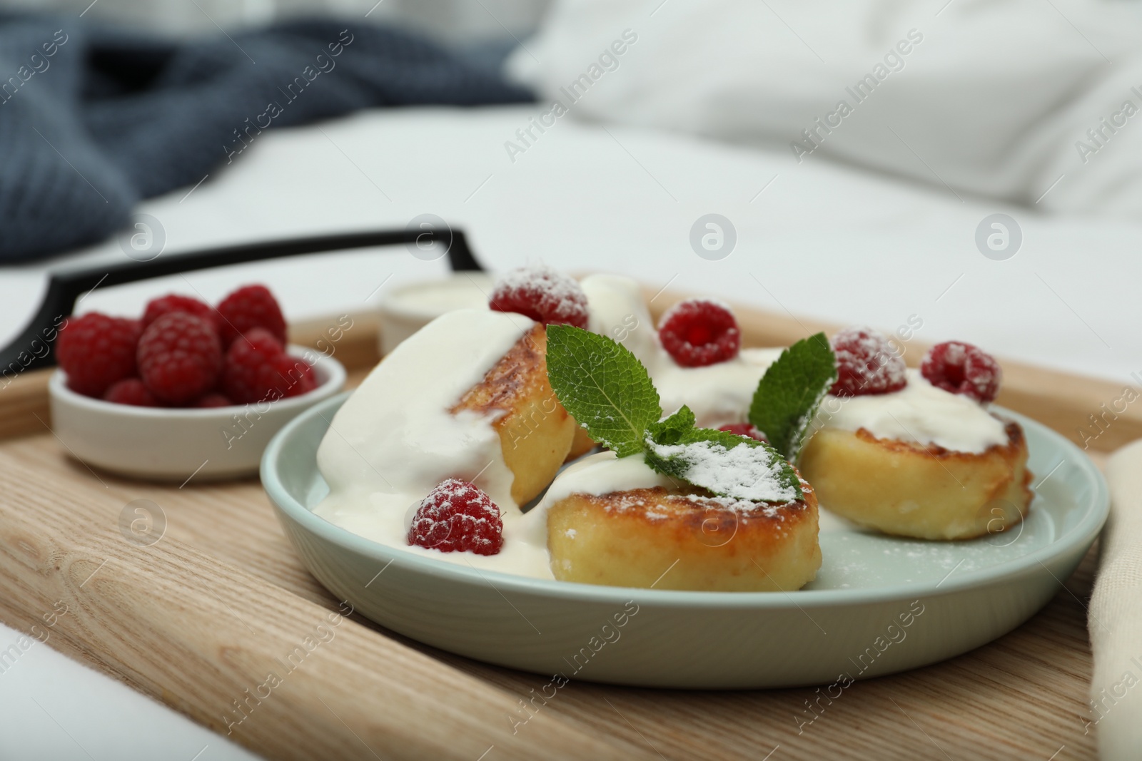 Photo of Tasty breakfast served in bedroom. Cottage cheese pancakes with fresh raspberries, mint, sour cream and icing sugar on wooden tray, closeup