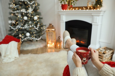 Photo of Woman with hot drink resting near fireplace in cozy room decorated for Christmas, closeup