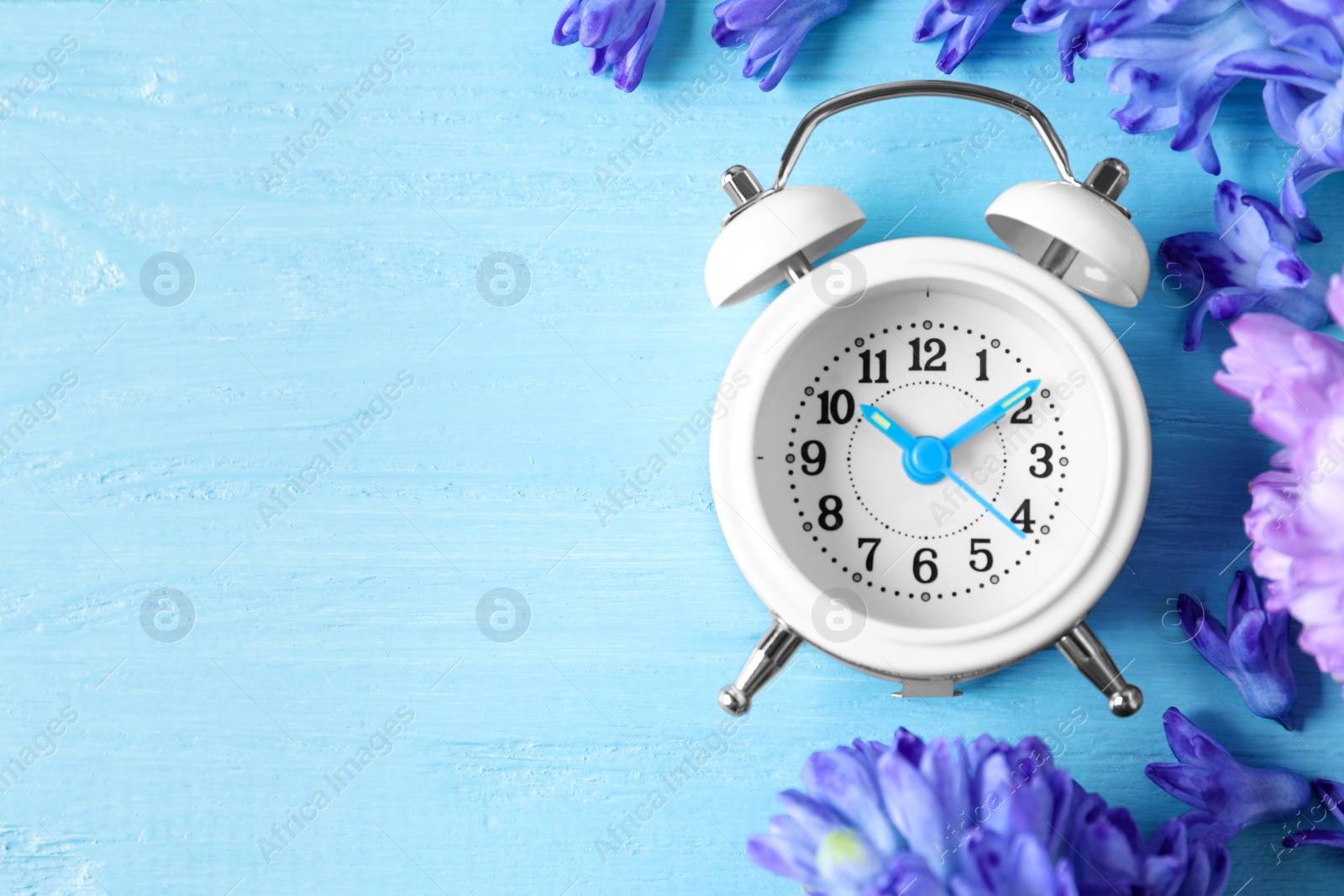 Photo of White alarm clock, spring flowers and space for text on light blue wooden table, flat lay. Time change
