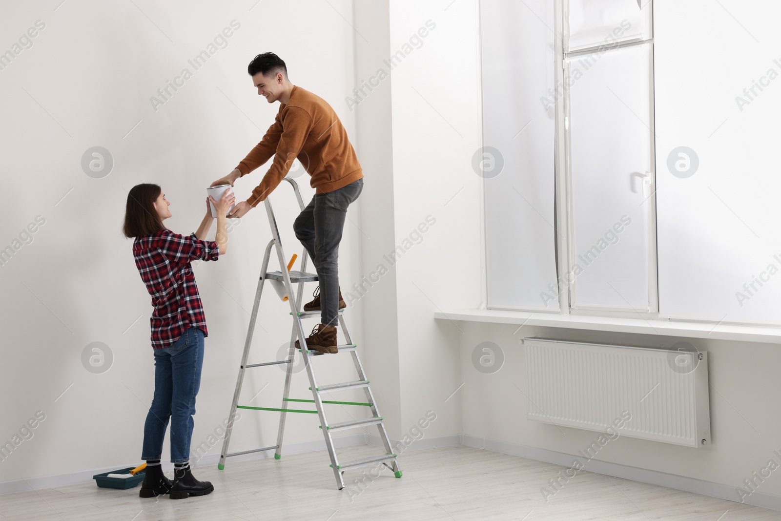 Photo of Young woman giving paint can to boyfriend on stepladder indoors. Room renovation