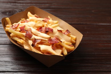 Tasty potato fries, cheese sauce and bacon in paper container on wooden table, closeup