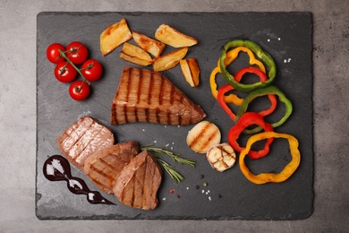 Photo of Slate plate with grilled meat, garnish and sauce on grey background, top view