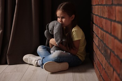 Child abuse. Upset little girl with toy bunny sitting on floor near brick wall indoors