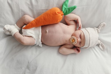 Adorable little baby with pacifier and carrot toy sleeping on bed, top view