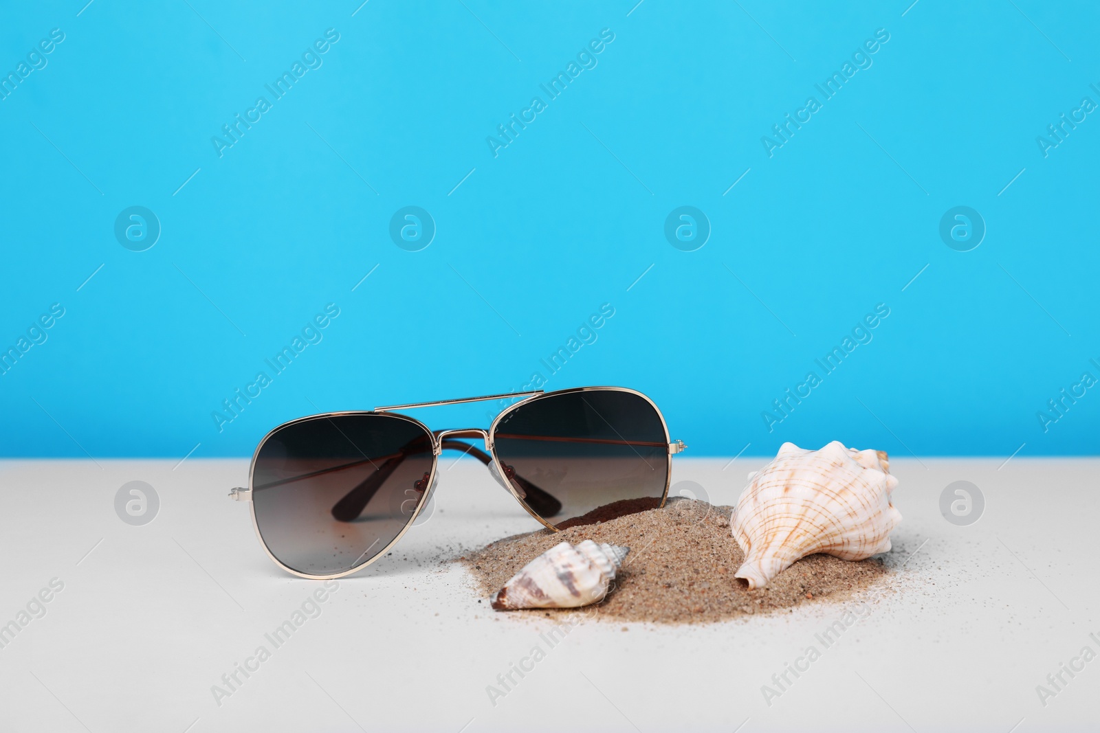 Photo of Stylish sunglasses, seashells and sand on white table against light blue background. Space for text