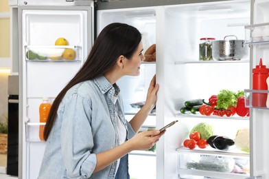 Photo of Young woman with smartphone near modern refrigerator in kitchen