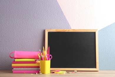 Photo of Different school stationery and small blank chalkboard on table near color wall. Space for text