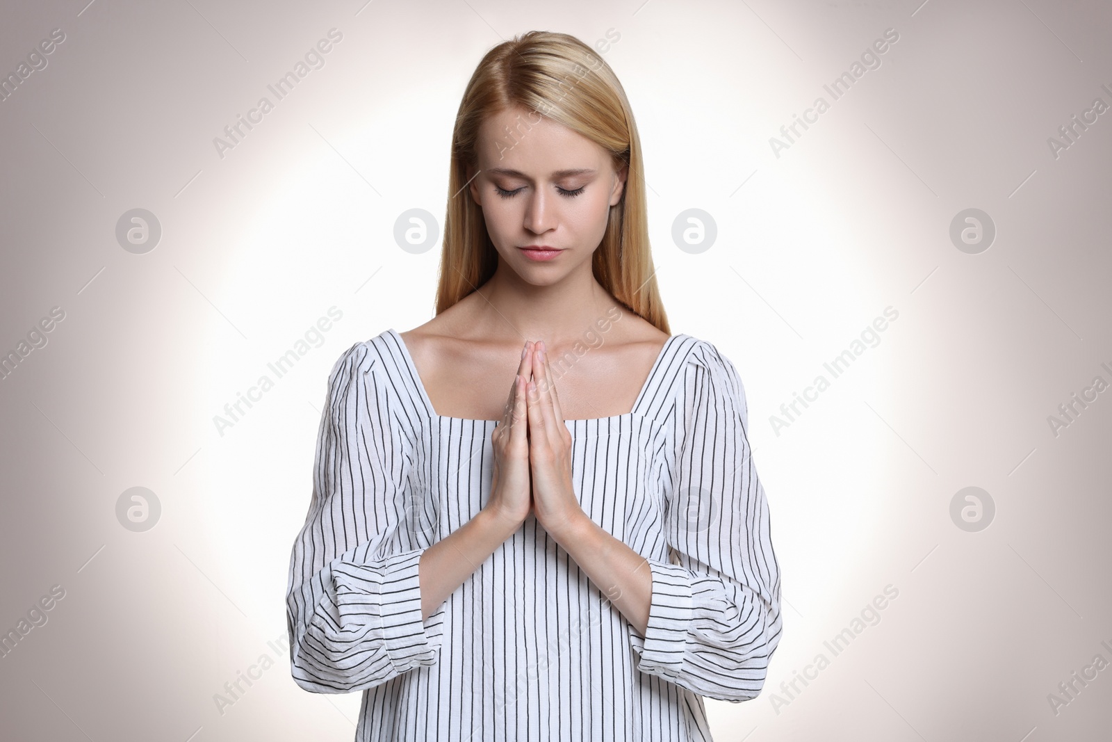 Photo of Religious young woman with clasped hands praying against light background