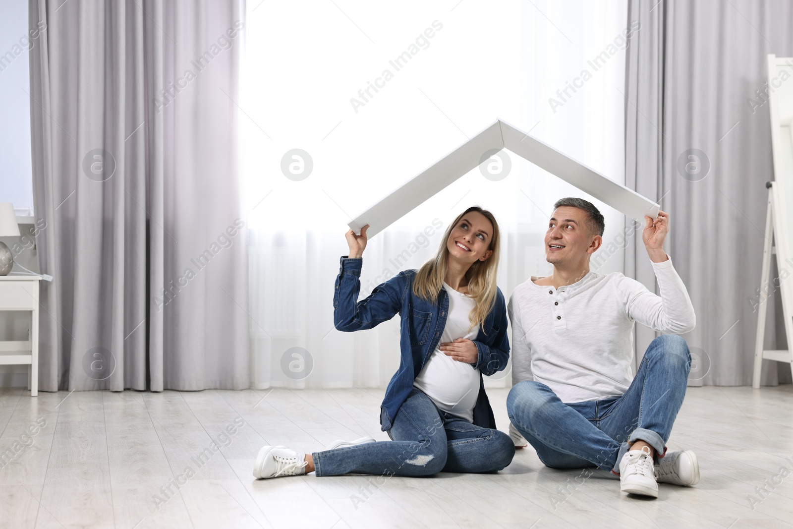 Photo of Young family housing concept. Pregnant woman with her husband sitting under cardboard roof on floor indoors. Space for text