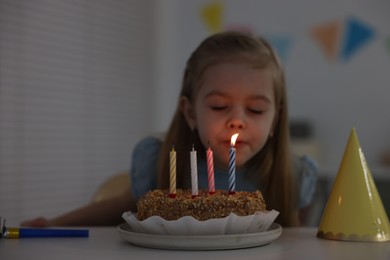 Photo of Cute girl blowing out birthday candles at table indoors, focus on cake