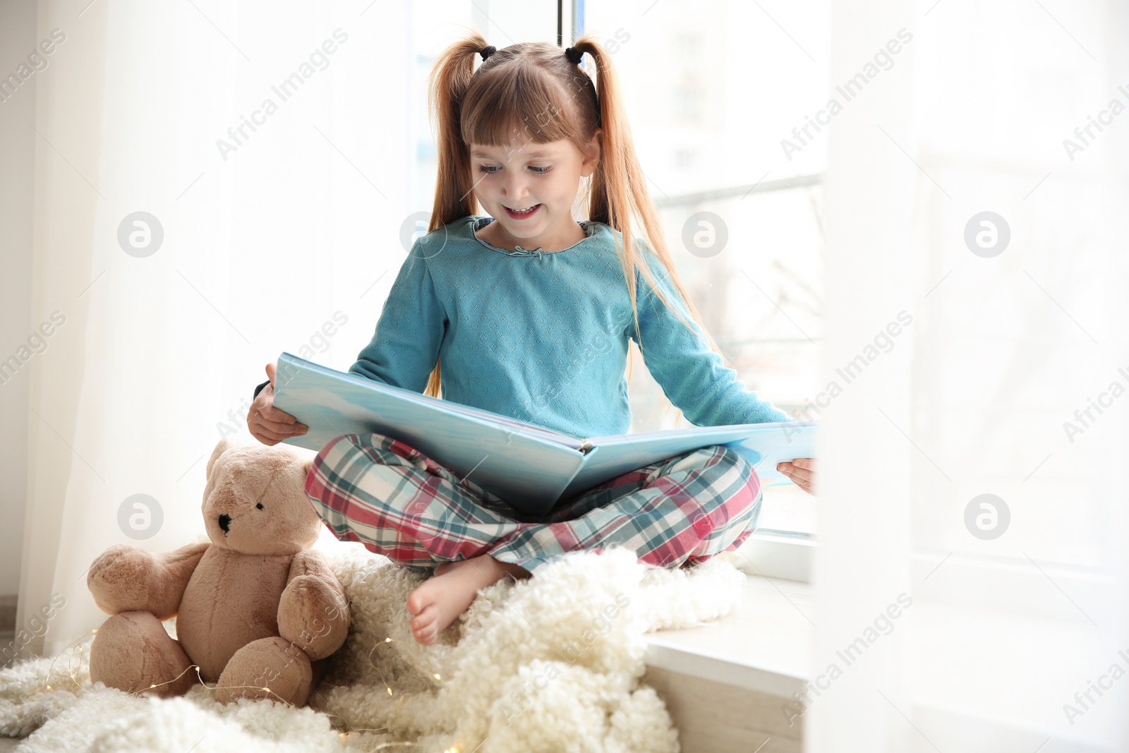 Photo of Cute little girl reading book near window at home