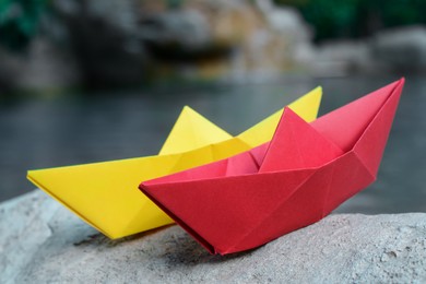 Photo of Beautiful yellow and red paper boats on stone near pond, closeup