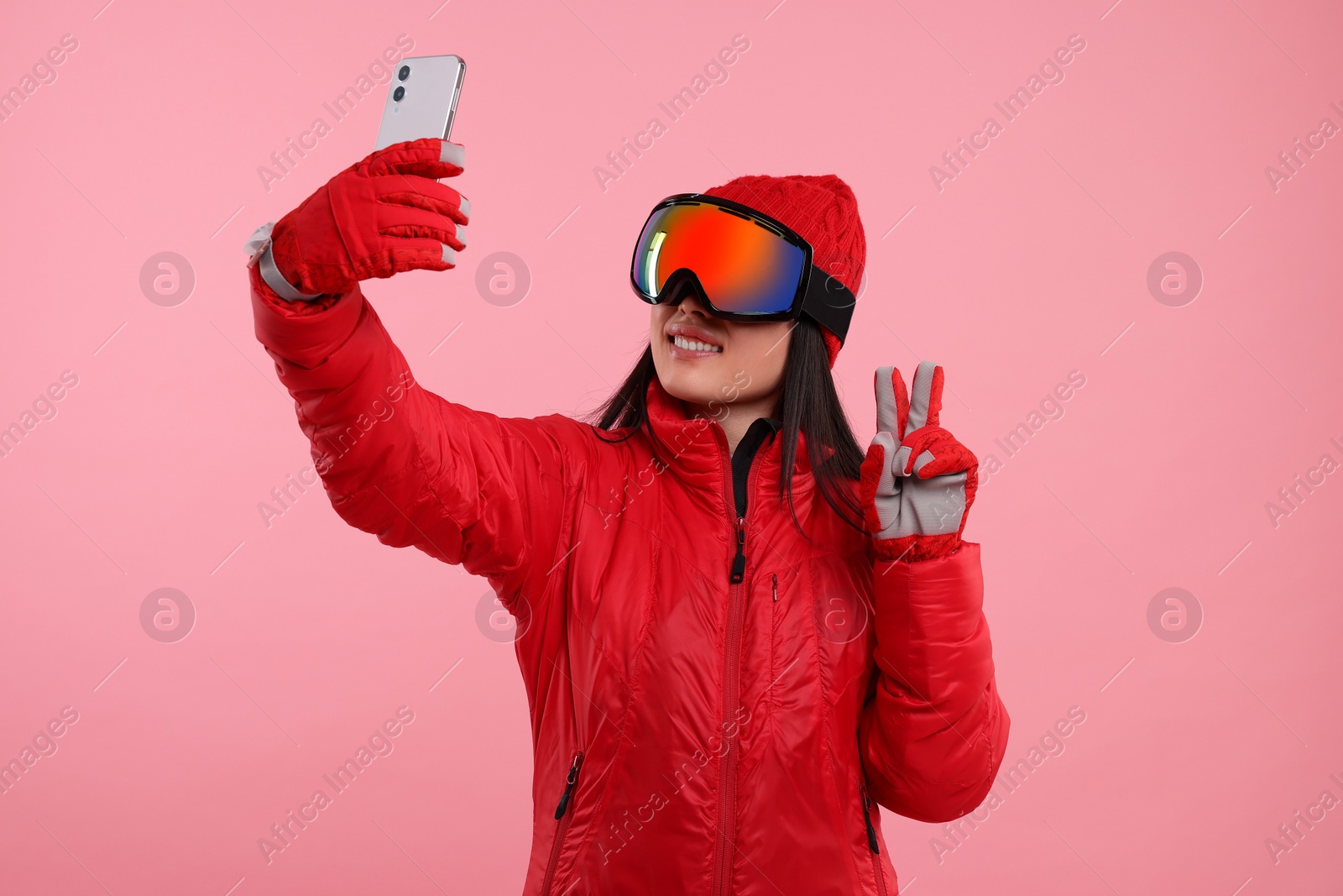 Photo of Happy woman in winter sportswear and goggles taking selfie and showing peace sign on pink background