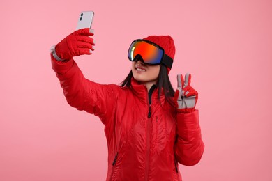 Photo of Happy woman in winter sportswear and goggles taking selfie and showing peace sign on pink background
