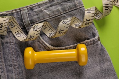 Jeans, dumbbell and measuring tape on green background, flat lay. Weight loss concept