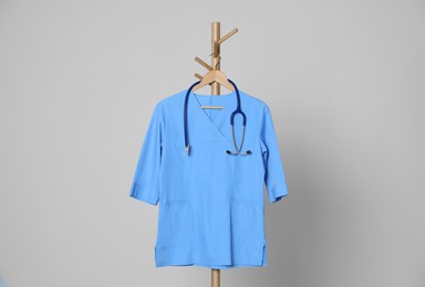 Medical uniform and stethoscope hanging on rack against light grey background