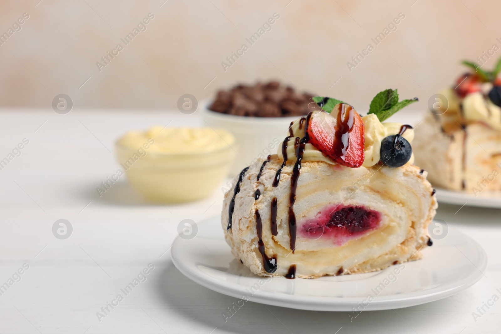 Photo of Piece of tasty meringue roll with jam, cream, strawberry, blueberry and mint on white wooden table, closeup. Space for text
