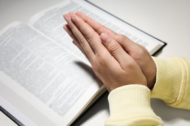 Photo of Woman holding hands clasped while praying over Bible at white table, closeup