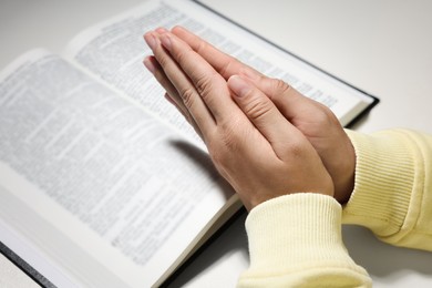 Woman holding hands clasped while praying over Bible at white table, closeup