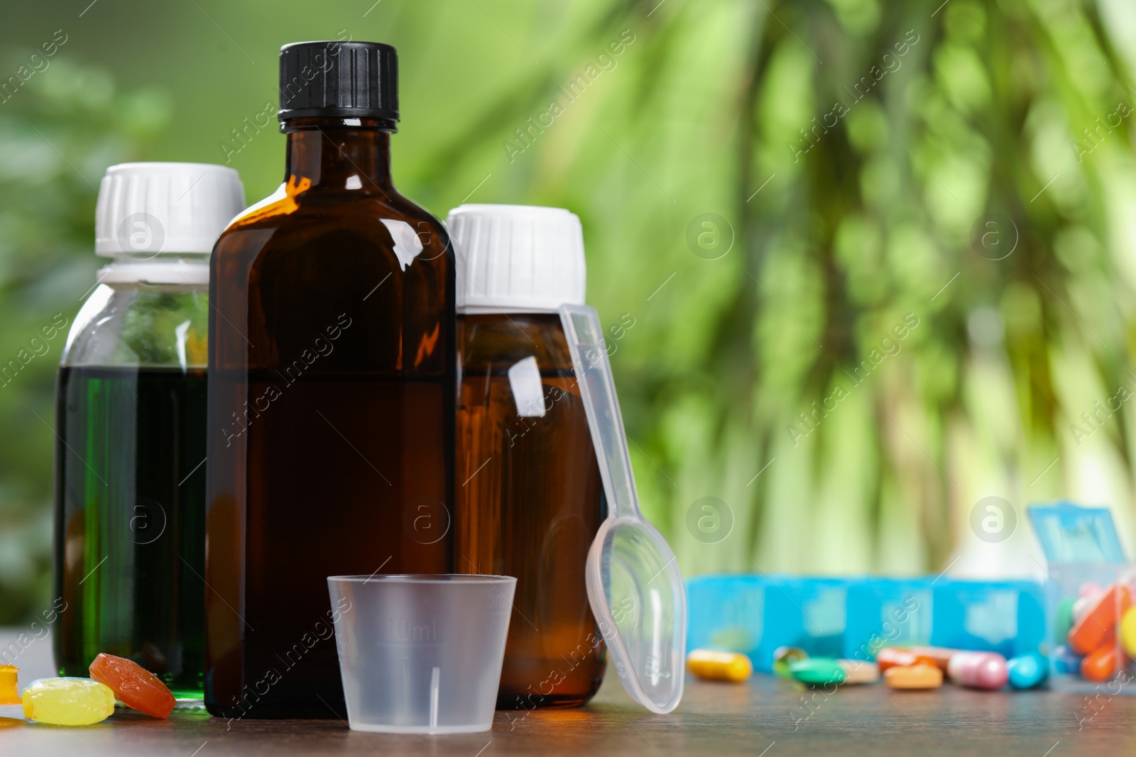 Photo of Bottles of syrup, measuring cup, dosing spoon and pills on wooden table against blurred background, space for text. Cold medicine