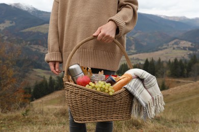 Woman holding wicker picnic basket with thermos, snacks and plaid in mountains on autumn day, closeup