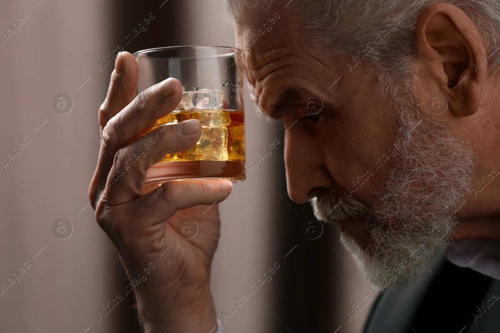 Photo of Senior man holding glass of whiskey with ice cubes on brown background, closeup