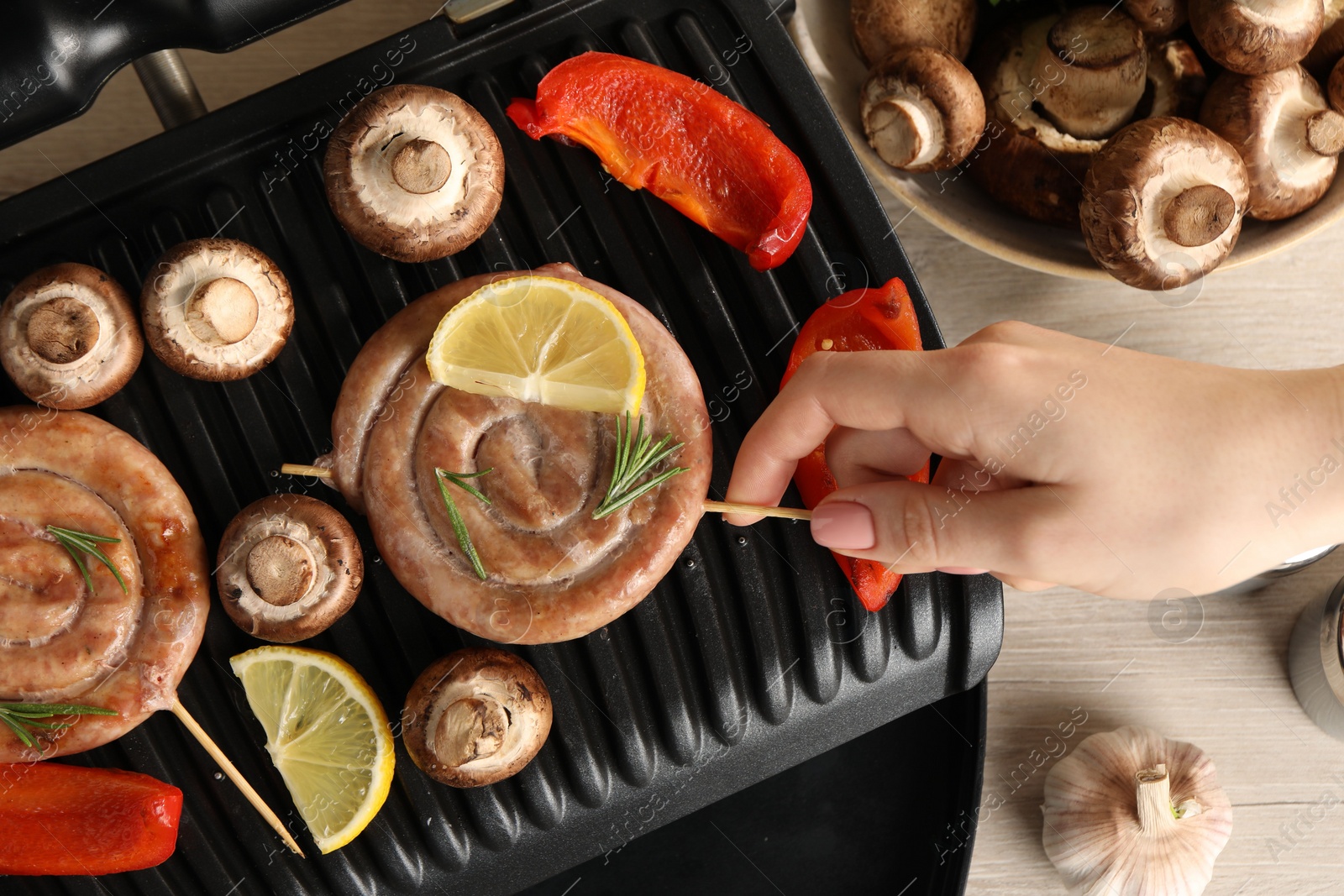 Photo of Woman cooking homemade sausages with mushrooms and bell pepper on electric grill at wooden table, top view