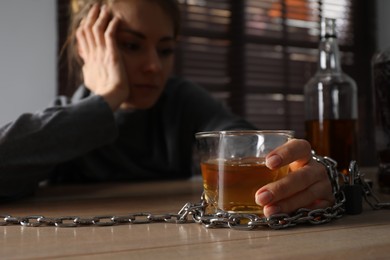 Photo of Alcohol addiction. Woman chained with glass of liquor at wooden table in room, focus on hand