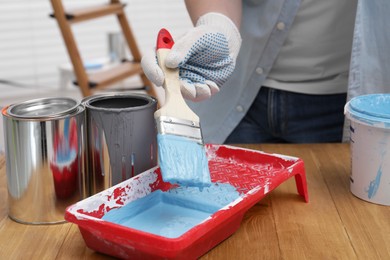 Photo of Man taking light blue paint with brush from tray at wooden table indoors, closeup