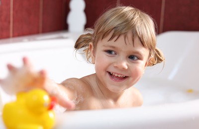 Photo of Smiling girl taking toy duck in bathtub at home, selective focus