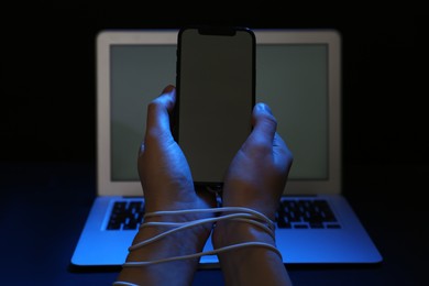 Photo of Woman with tied hands holding smartphone near laptop on dark background, closeup. Internet addiction