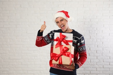 Photo of Happy man in Christmas sweater and Santa hat holding gift boxes near white brick wall