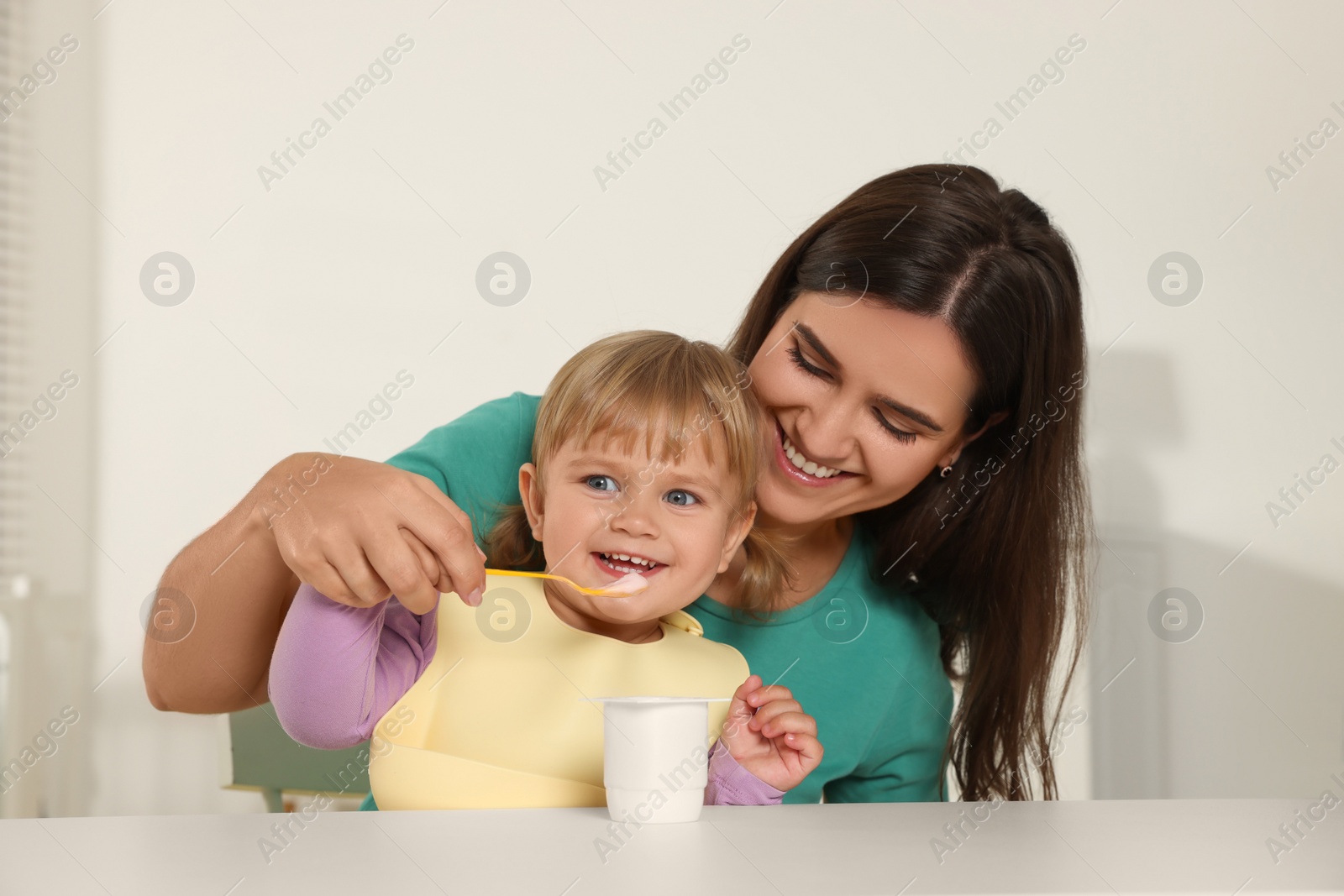 Photo of Mother feeding her cute little child with yogurt at white table in room