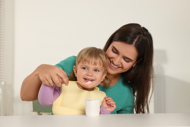 Mother feeding her cute little child with yogurt at white table in room