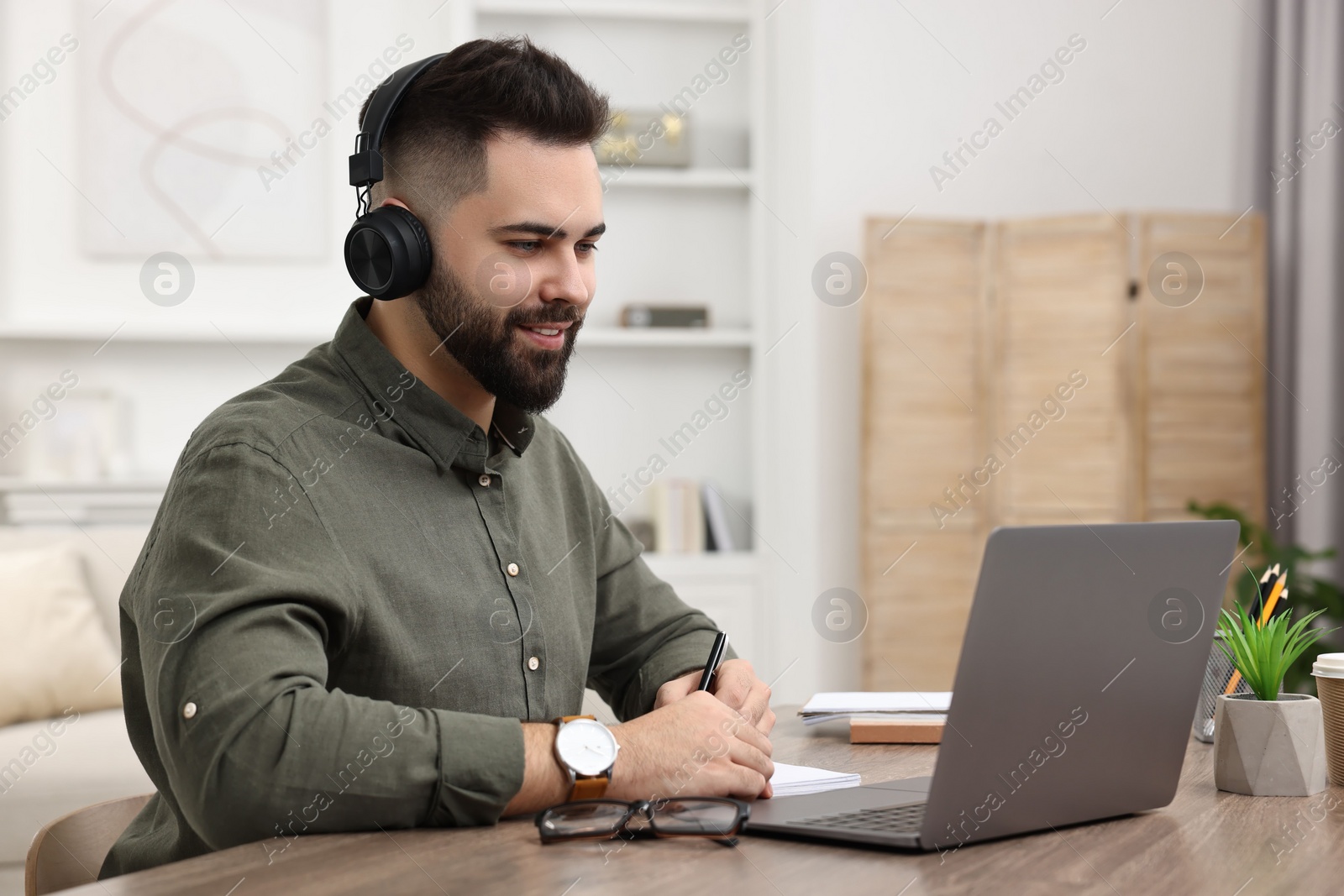 Photo of E-learning. Young man taking notes during online lesson at wooden table indoors