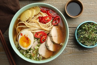 Photo of Delicious ramen in bowl, soy sauce and microgreens on wooden table, flat lay. Noodle soup