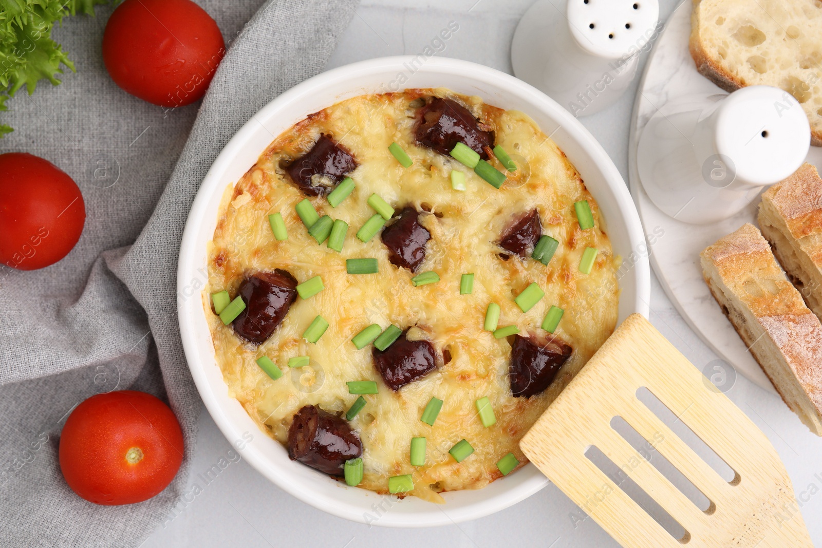 Photo of Tasty sausage casserole with green onions in baking dish served on white tiled table, flat lay